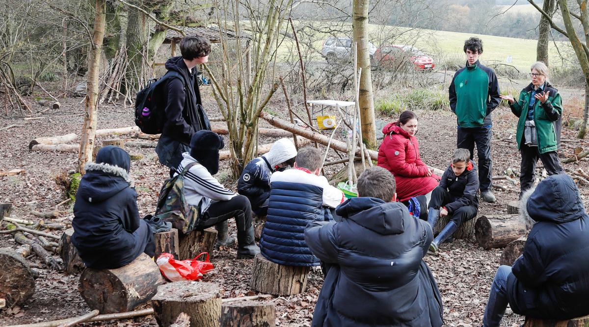 A group of schoolchildren sat on logs whilst listening to two members of staff giving a brief