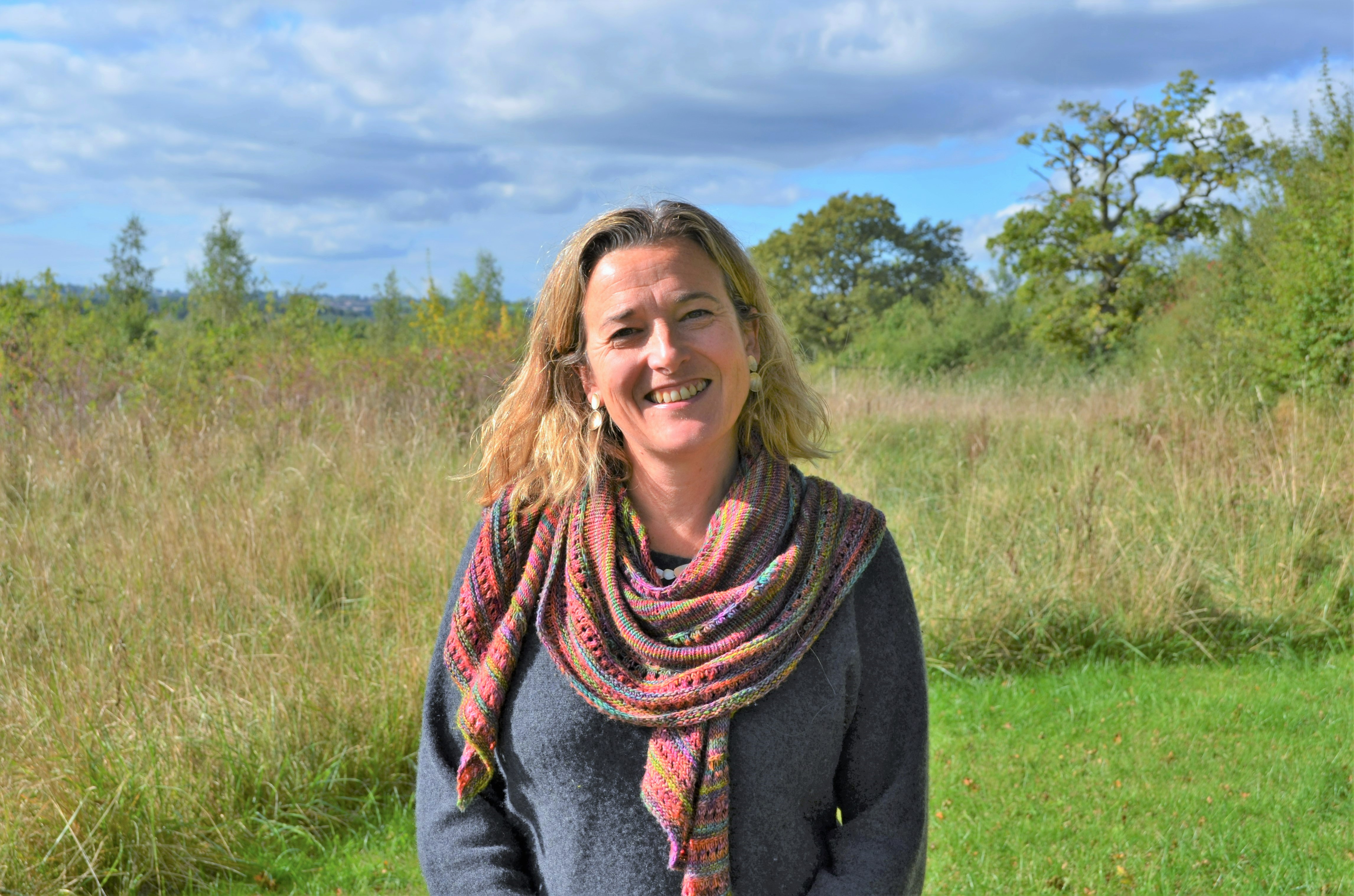Nicky smiling in a colourful scarf in front of a green wildflower meadow