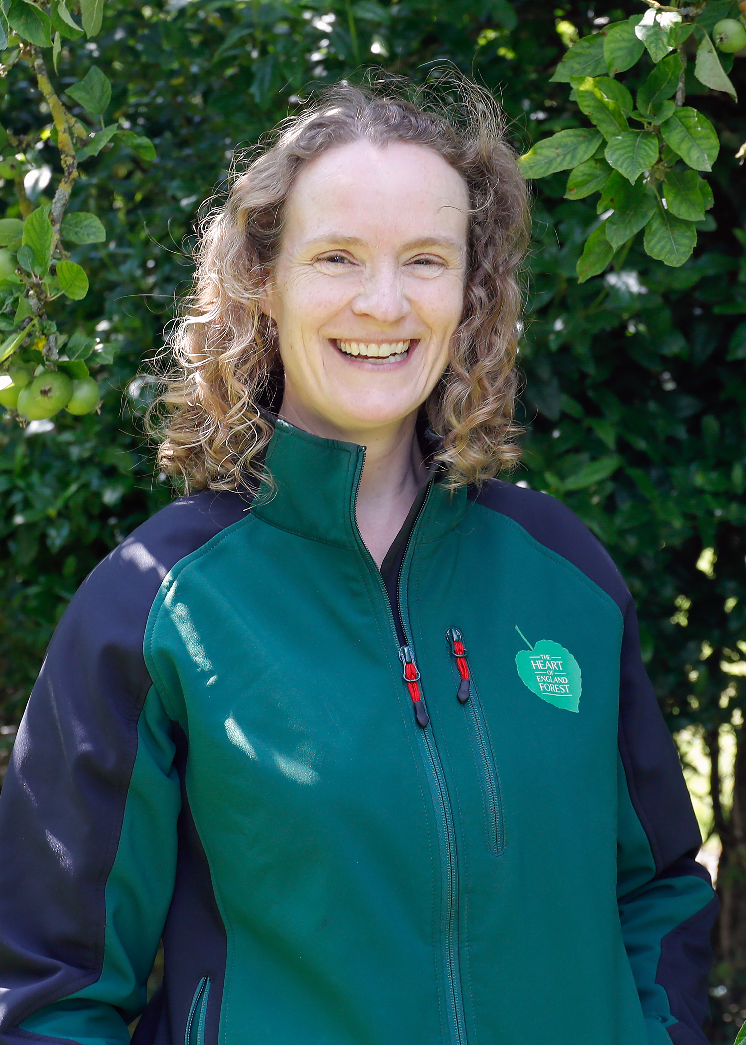 Close up portrait photo of Sophie, our Biodiversity Manager, stood smiling in front of some green leaves and wearing a green Heart of England Forest jacket.