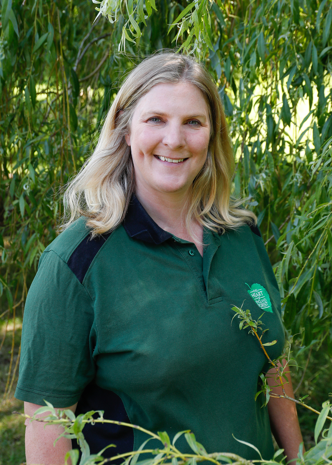 Close up of Elaine, our Head of Learning and Skills, stood smiling, surrounded by green leaves and wearing a branded, green Heart of England polo shirt.