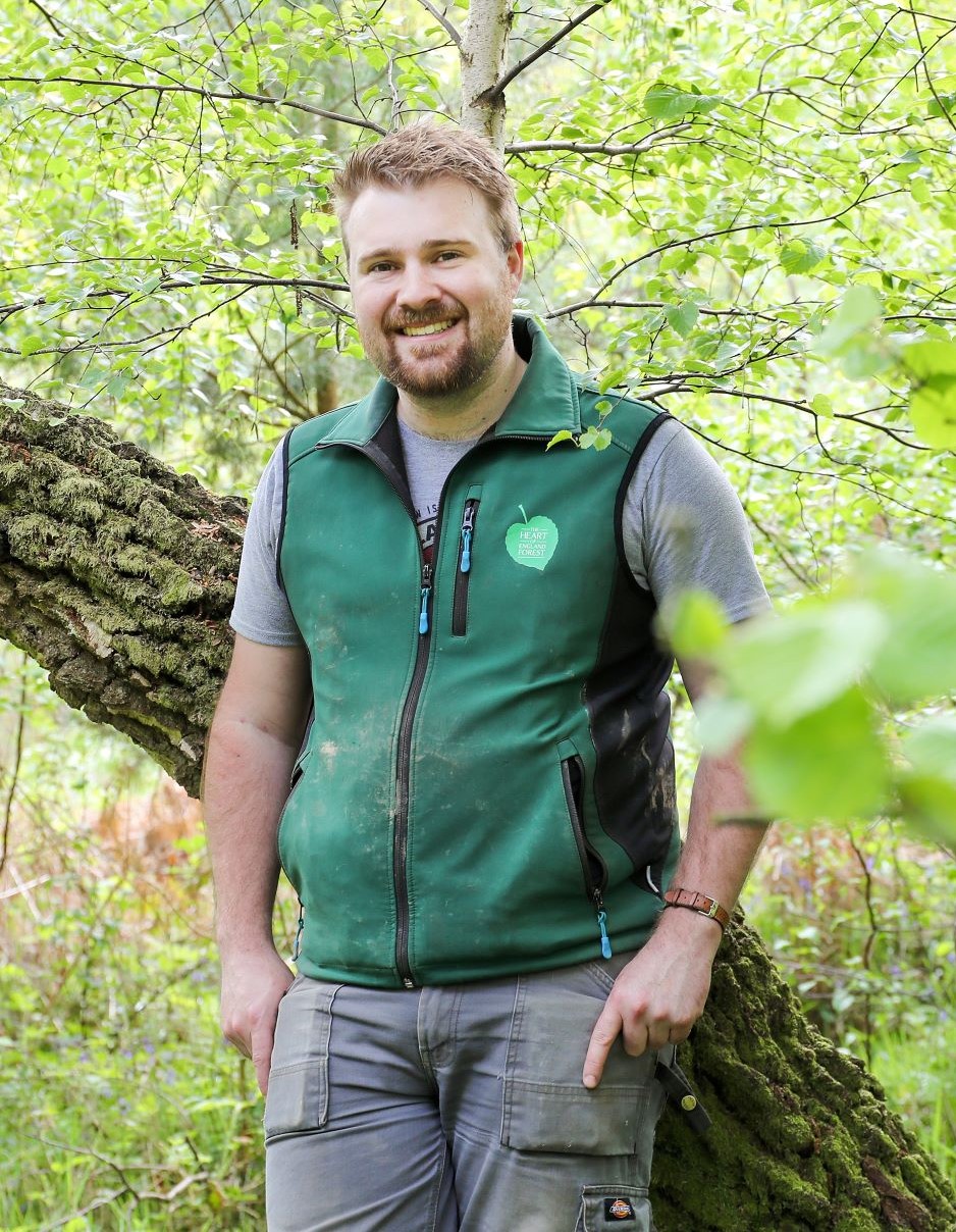 Jonathan leaning against a tree trunk wearing a Heart of England Forest branded gilet and smiling at the camera 