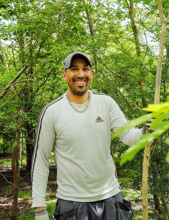 Male volunteer Leon standing in the Forest holding on to a thin tree trunk. He is smiling at the camera and there are green leaves in the foreground.