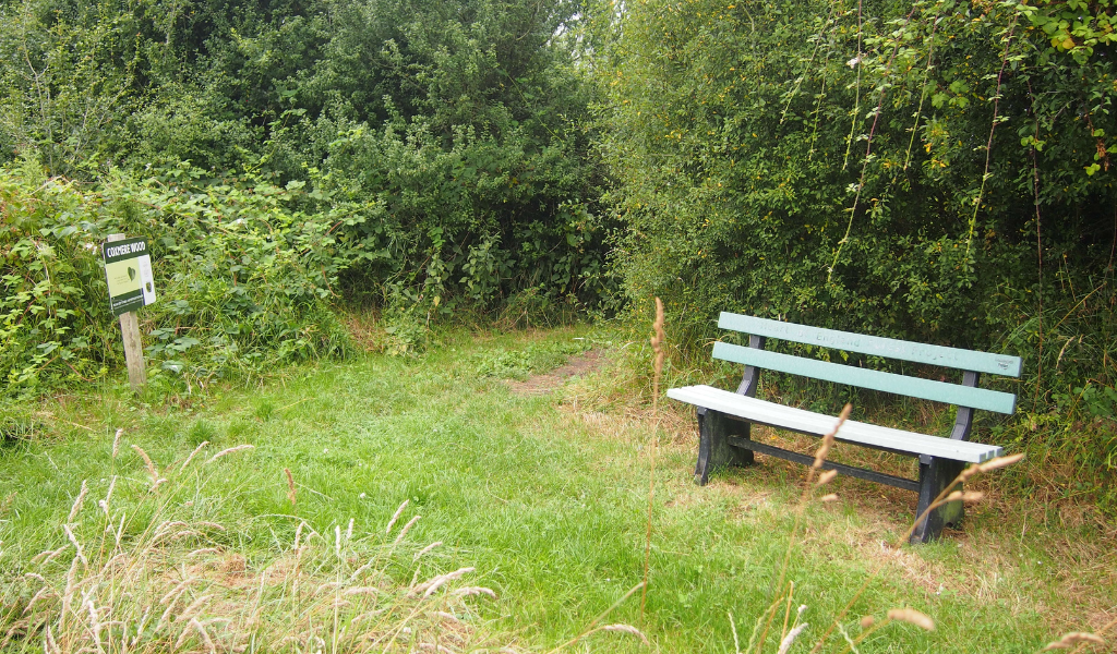A bench on the right and a 'Coxmere Wood' sign on the left along a woodland path with mature trees surrounding