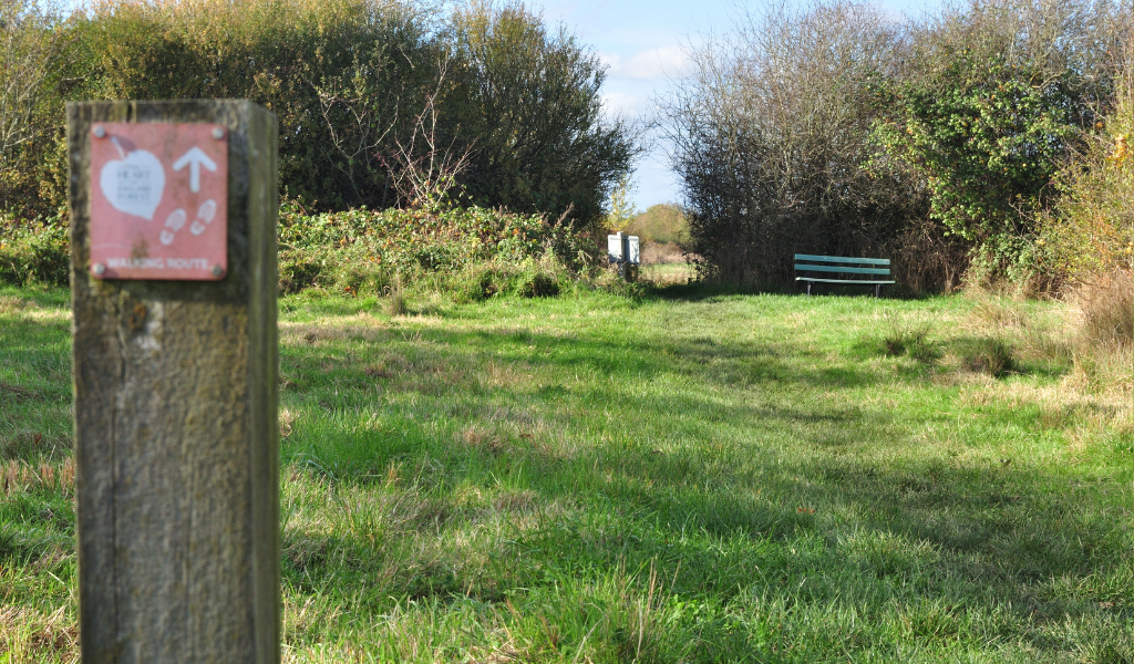 A waymarker post in the left hand corner directing the path towards a gap in the hedge ahead with a bench and a Coxmere Wood sign next to it