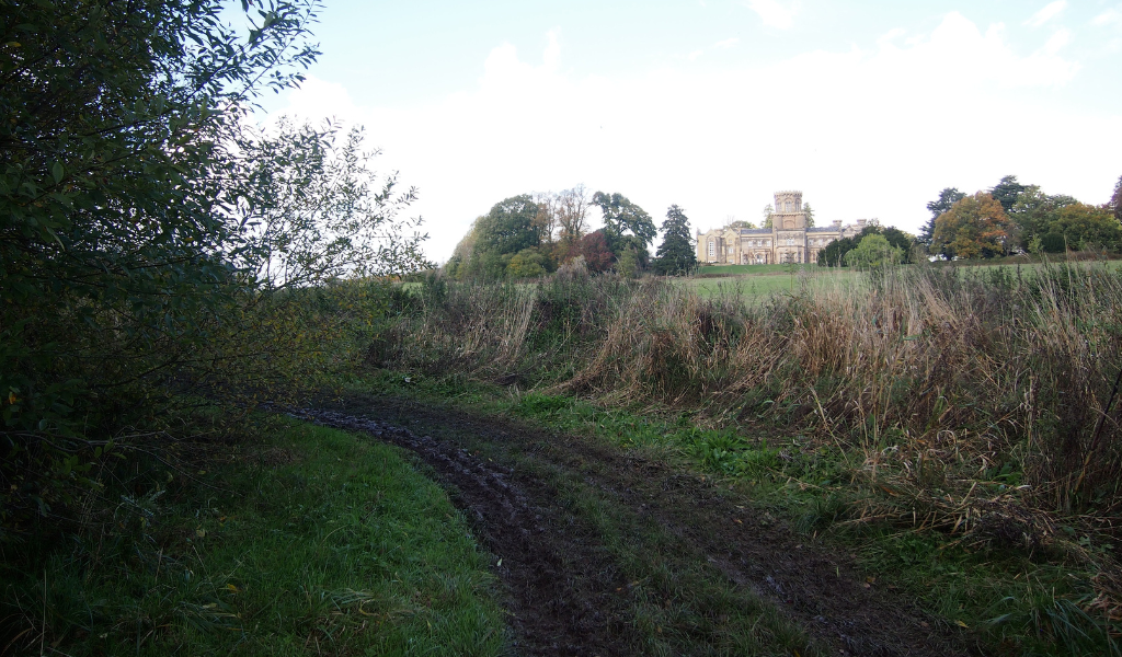 Studley Castle in the distance on the right with an open field in front of it. There is a mature hedgerow running along the left hand side.