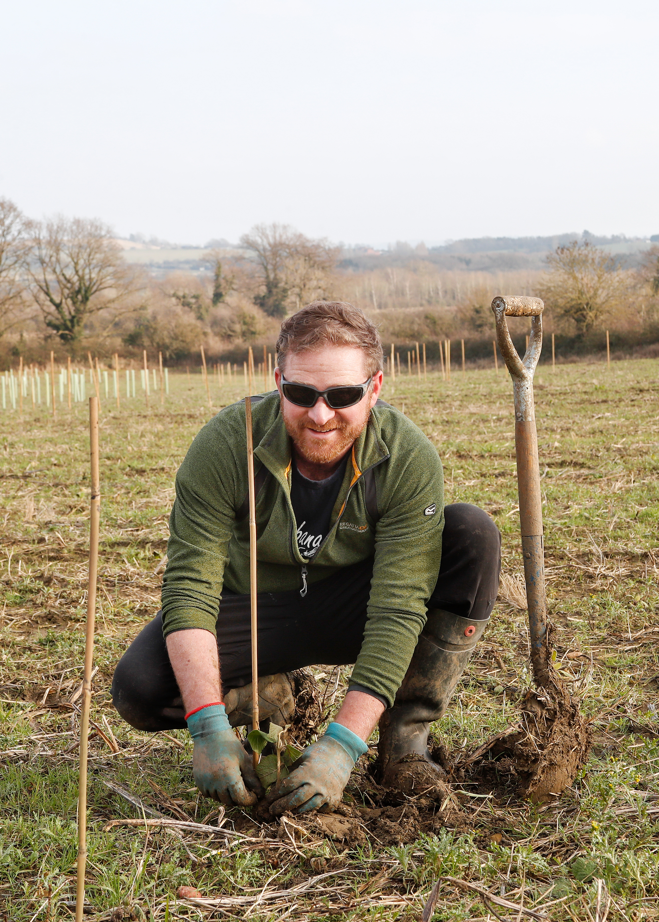 A photograph of Ian Hayward wearing sunglasses, planting a tree and smiling at the camera