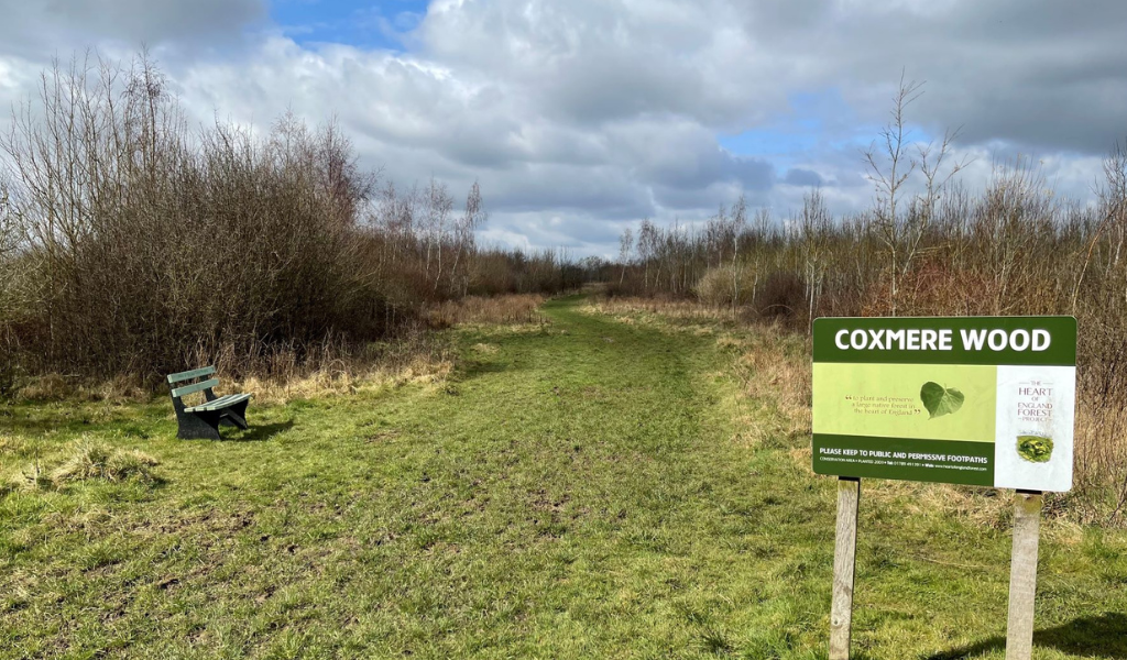 A bench to the left and a Coxmere Wood sign to the right along the footpath