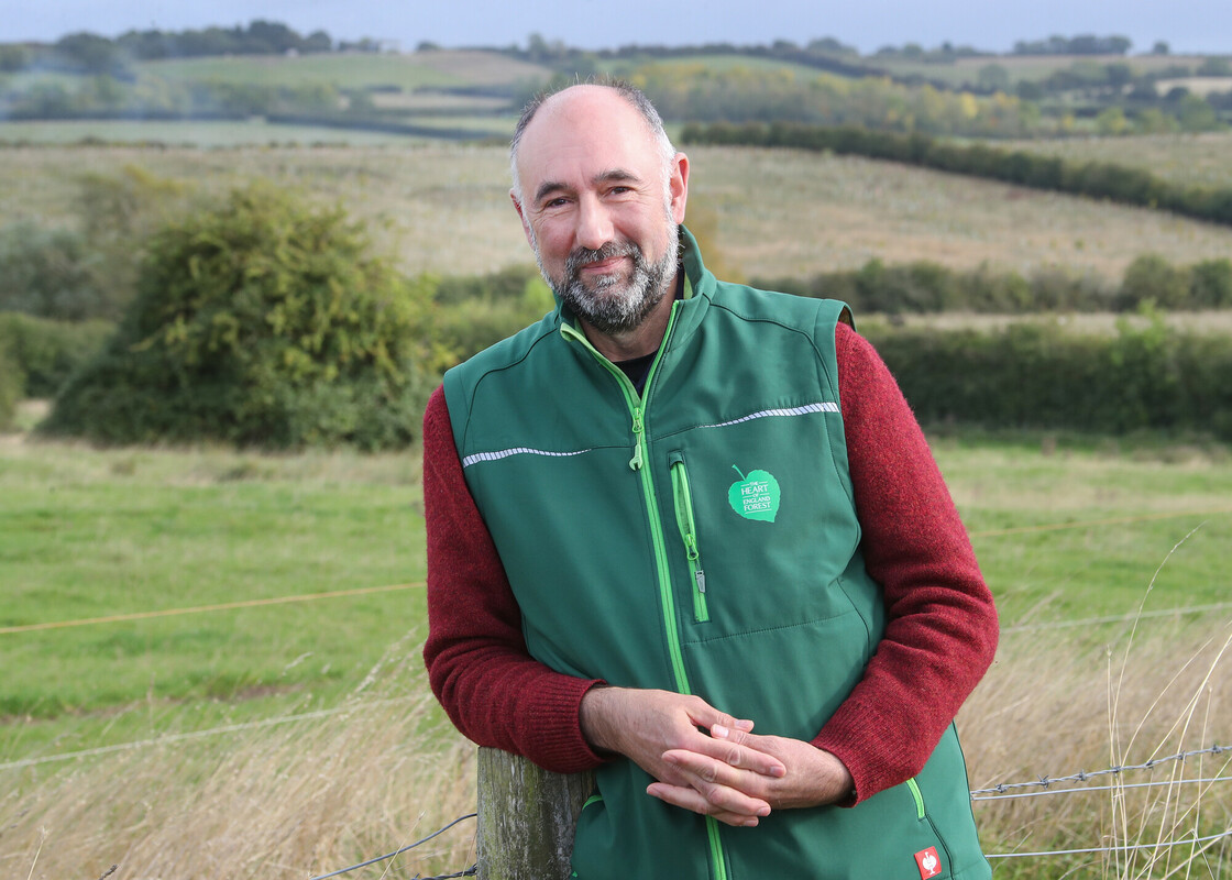 Ed Leszczynski  leaning on a post looking at the camera, with rolling hills in the background