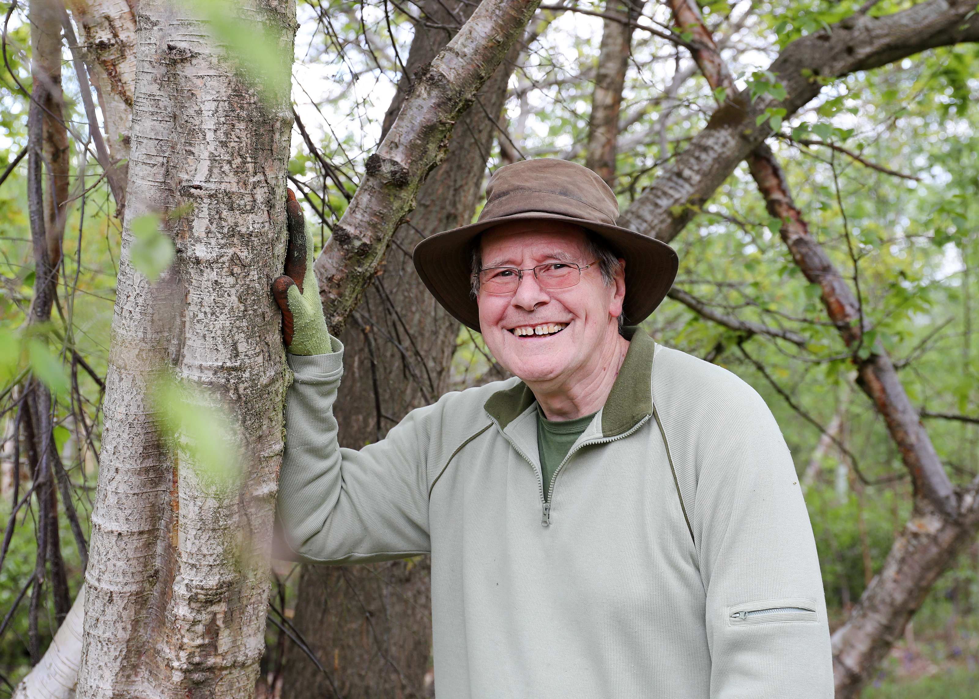 Close up of Alan smiling and wearing a hat leaning against a tree branch