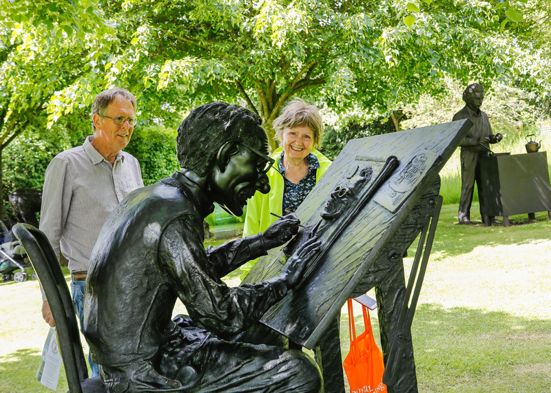 A male and a female visitor standing smiling at a bronze statue with another statue and leafy trees behind