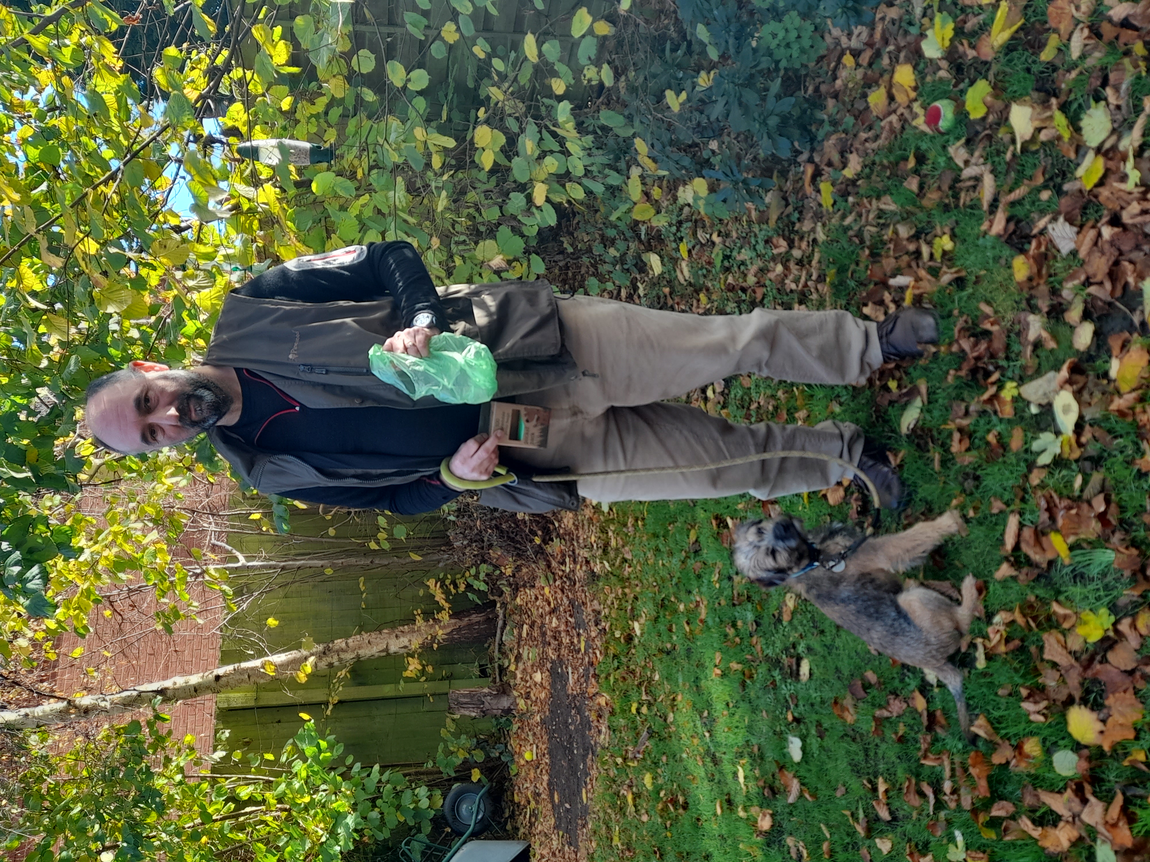Ed standing in a grassy garden full of fallen autumnal leaves holding a green sustainable poo bag in one had and his dog’s lead in the other. His puppy sits to his left.