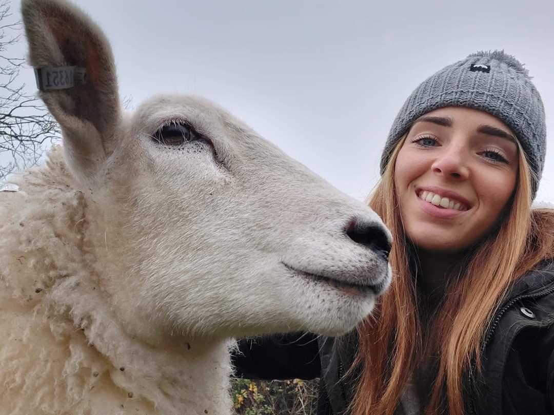 A selfie of Emma smiling wearing a grey beanie next to the white face of one of our lleyn sheep. 