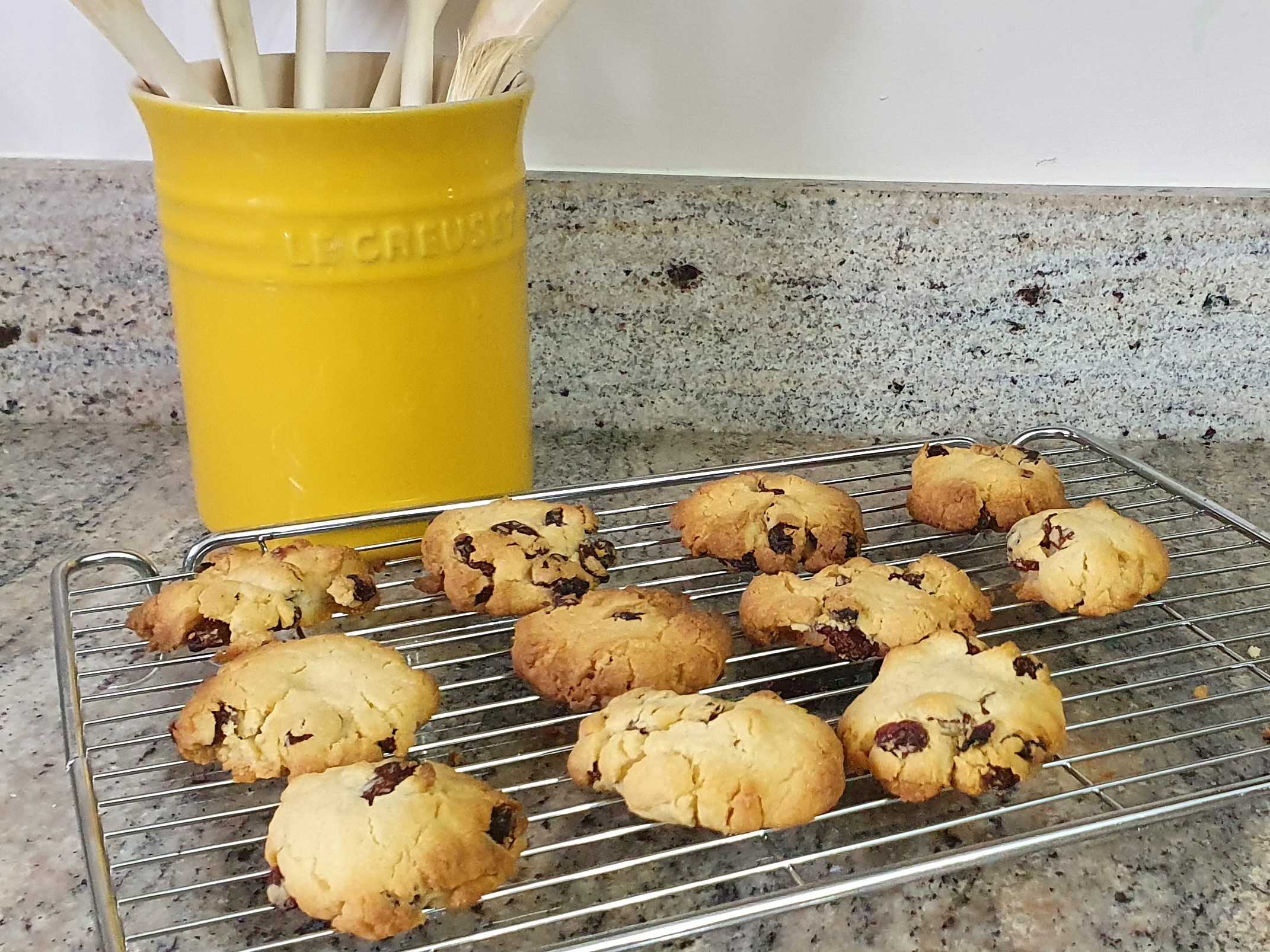 11 of Phil’s homemade cookies sitting on a cooling tray on a grey worktop with a yellow pot of wooden spoons behind them to the left.