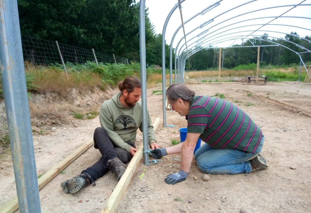 Two volunteers sitting on the ground helping to construct the metal frame for a polytunnel in our tree nursery