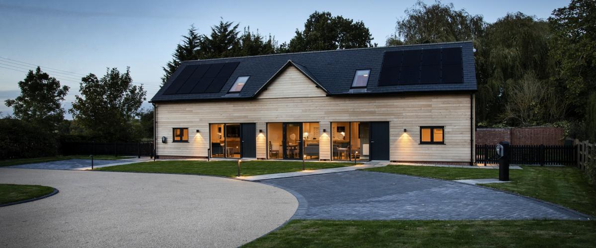 The exterior of both cottages at dusk. Driveway sits to the left with electric car charging points. Solar panels are fitted on the black roof.