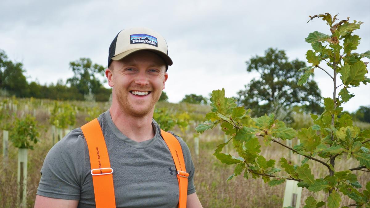 Aaron wearing a grey t-shirt, orange braces and a cap stood smiling in a field full of young trees.