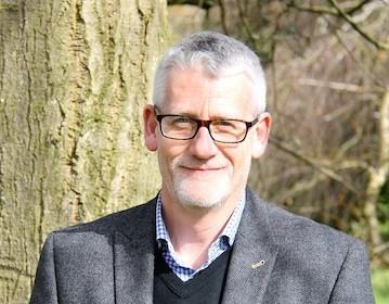 Close up of Steve wearing black glasses and a blazer in front of a tree trunk.