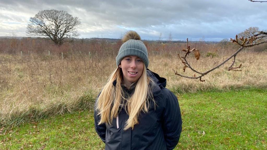 Close up of Ellie smiling in a fluffy grey beanie hat stood on some grass in the Forest.