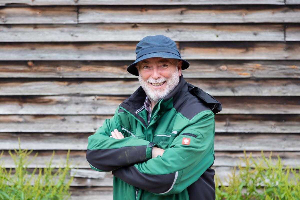 Phil standing with his arms crossed in front of a wooden panelled wall wearing a black bucket hat and a green branded jacket.