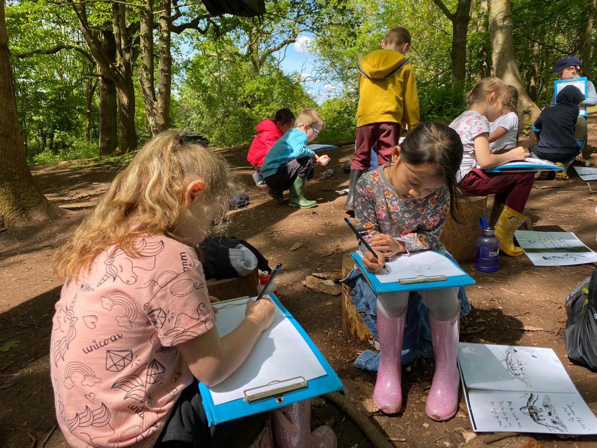 Two girls sitting on tree stumps writing on clipboards with classmates sitting behind