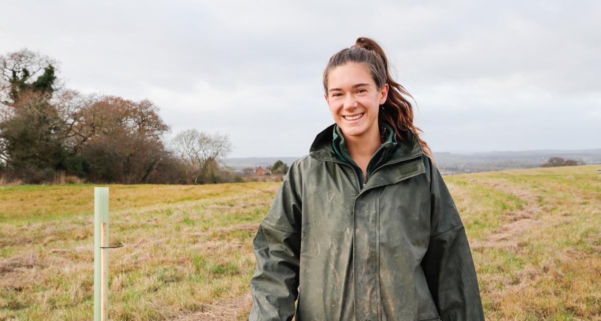 Forest Ranger Phoebe standing in a field smiling with her arm resting on a wooden tree stake, wearing a dark green waterproof coat, with newly planted trees in tubes behind her.