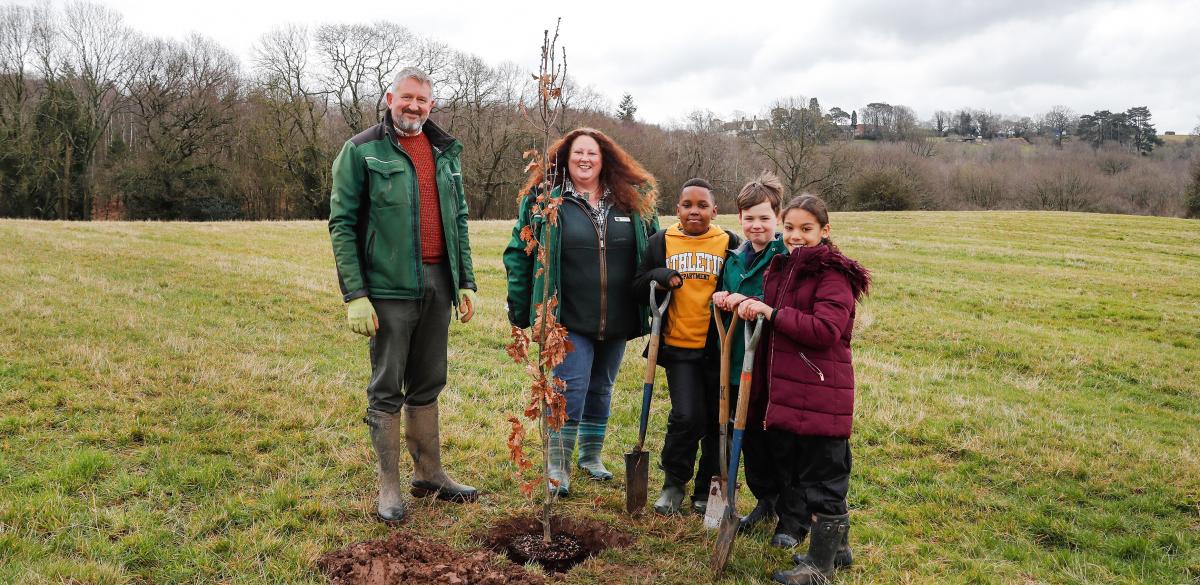 Head Forester Stephen, Chief Executive Beth Brook, and three school children all holding spades stand around an English oak tree sapling in a hole in the ground. They are all looking at the camera and smiling.