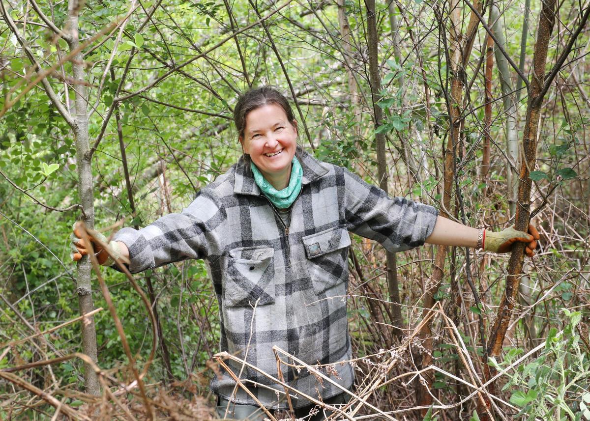 Female volunteer standing smiling at the camera holding onto two thin tree trunks either side of her and wearing gardening gloves in the Forest