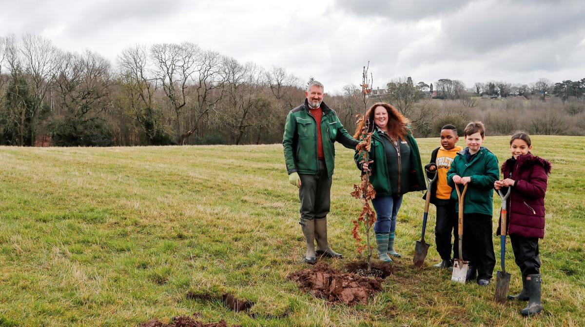 Our Head Forester and Chief Executive standing with three school pupils by a newly planted oak tree in a green field with mature trees in the distance