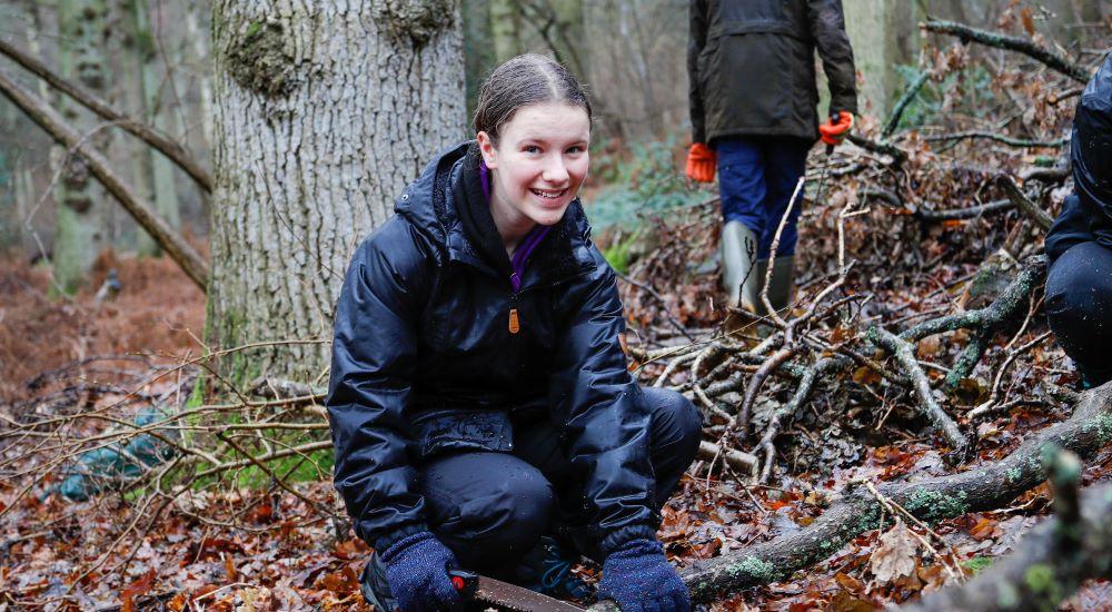 A teenage girl crouching on the forest floor wearing waterproof clothing. She is smiling at the camera