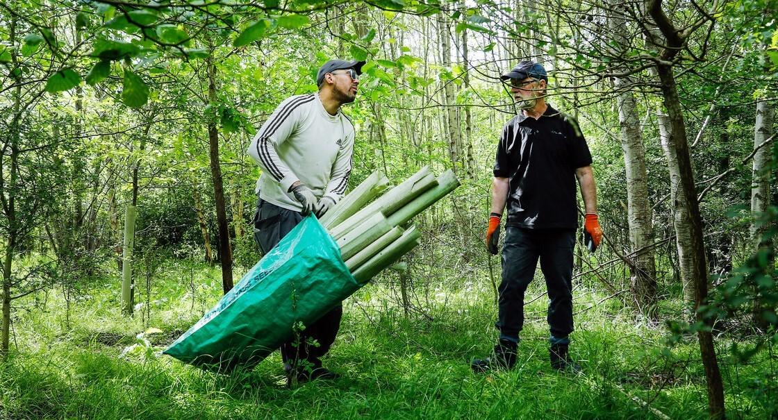 Two make volunteers standing in a Forest clearing, one has a sack of tree guards that have been removed