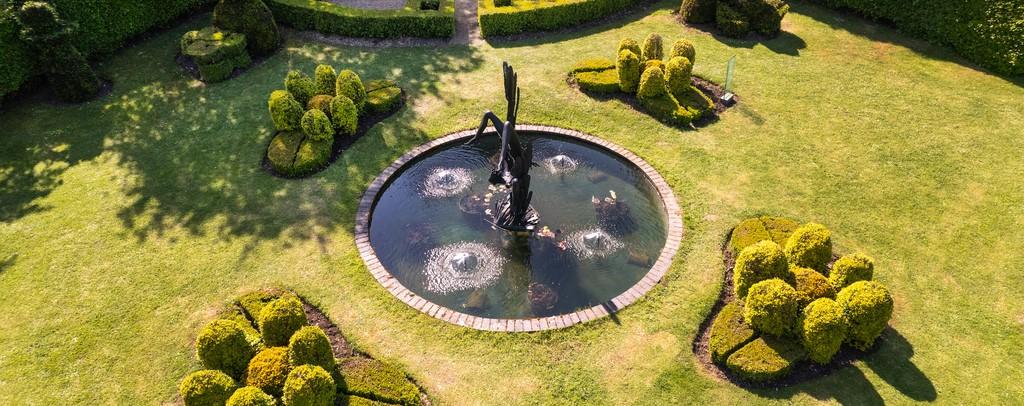 Aerial photo of landscaped lawn and bushes with a circular pond which has a bronze statue of Icarus in the middle