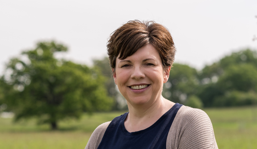 A head and shoulders photo of Alison Hunter taken with grass and trees in the background. Heather is smiling at the camera