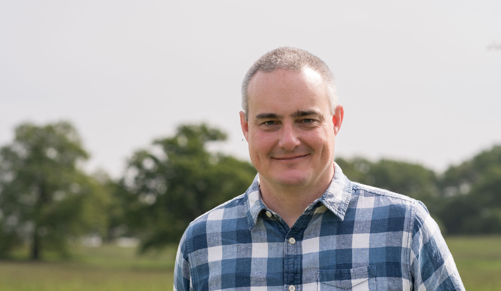 A head and shoulders photo of Asa Gurden taken with grass and trees in the background. Heather is smiling at the camera