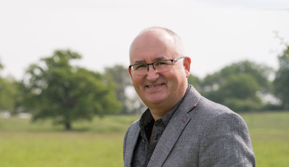 A head and shoulders photo of Dave Throup taken with grass and trees in the background. Heather is smiling at the camera