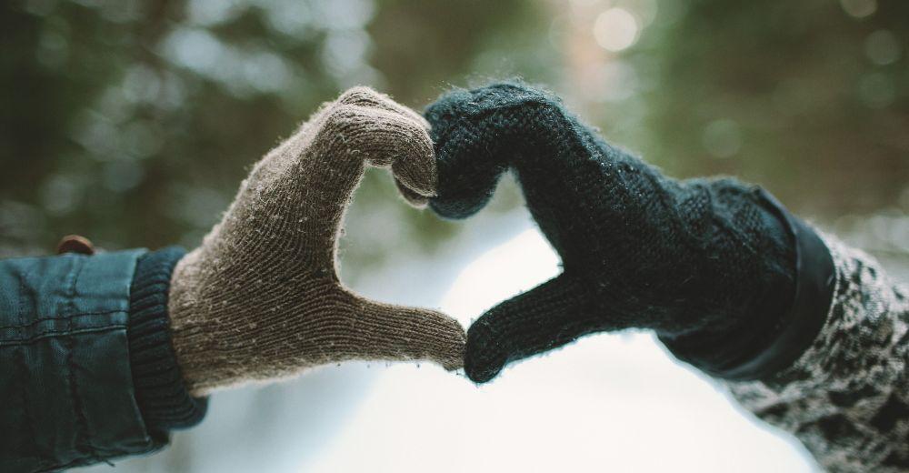 Two hands wearing woolly gloves forming a heart with a snowy forest in the background