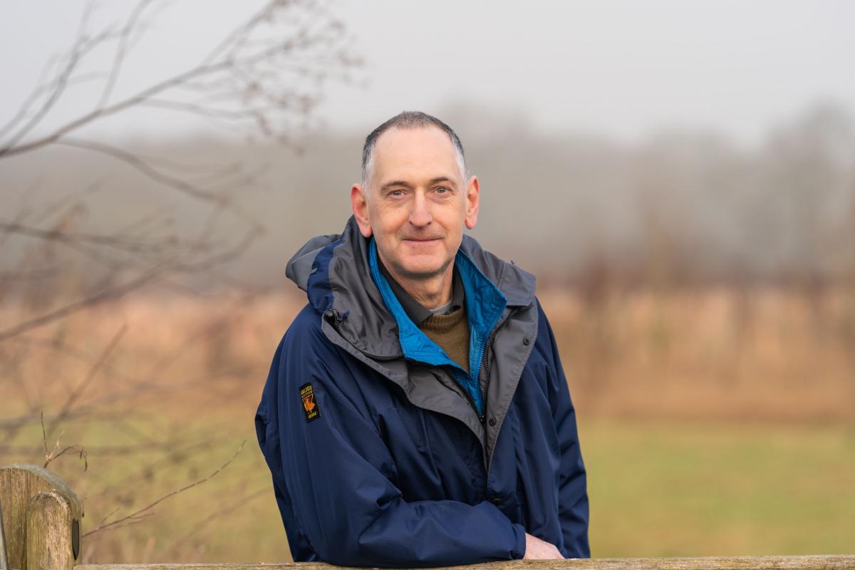 Rob Mackenzie, leaning on a post. He is smiling at the camera. This has been captured as a head and shoulders shot. 