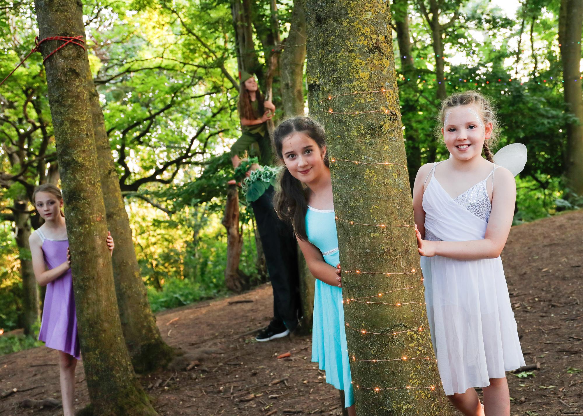 Three young girls in fairy costumes poking their smiling faces out from behind tree trunks