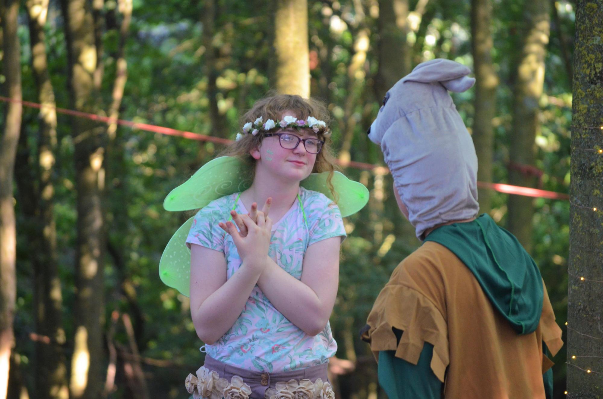 A young girl playing Titania wearing fairy wings and a flower crown clutching her hands to her heart.