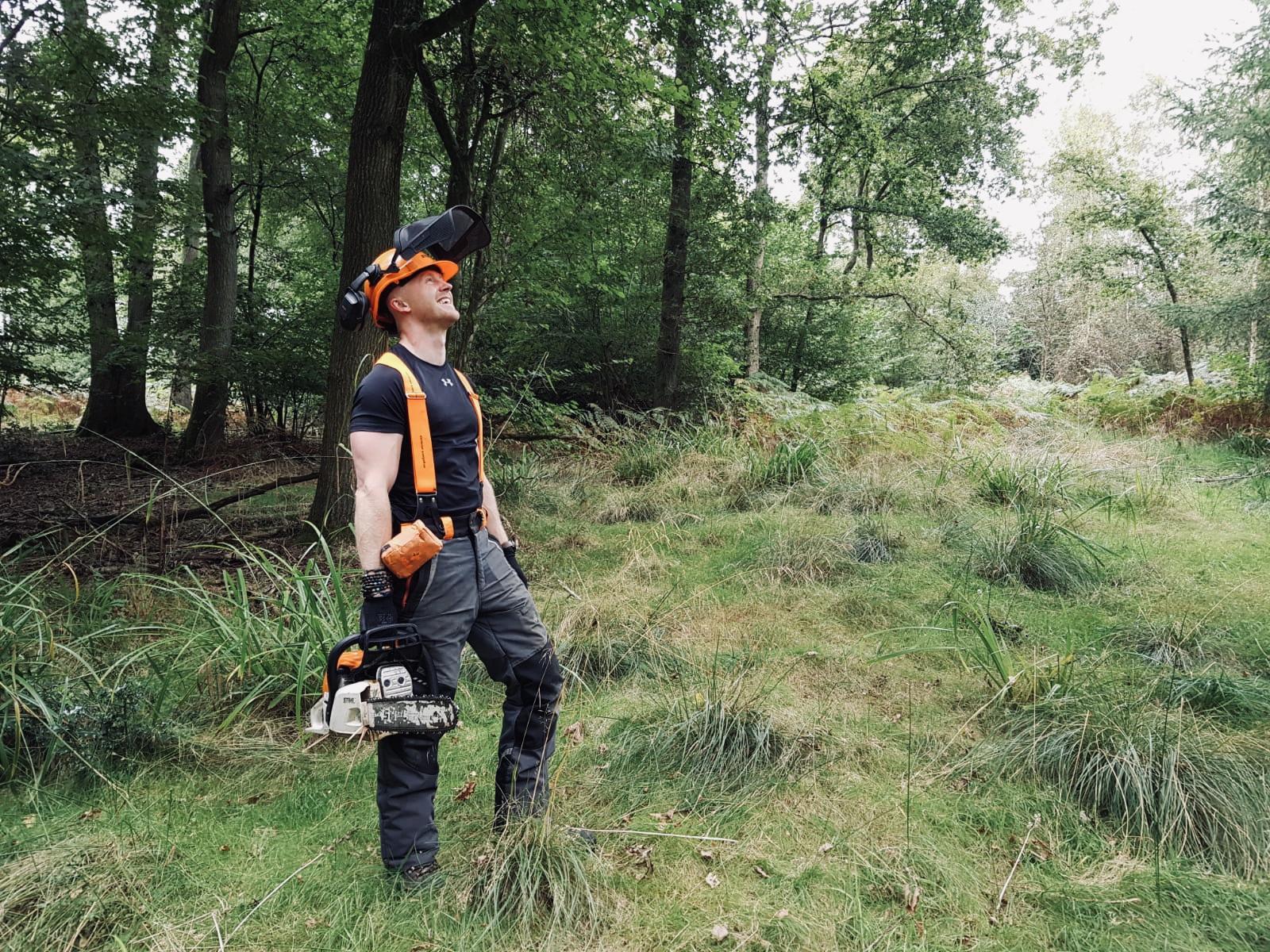 Aaron in protective gear and holding a saw, smiling and looking up into the tree canopy.