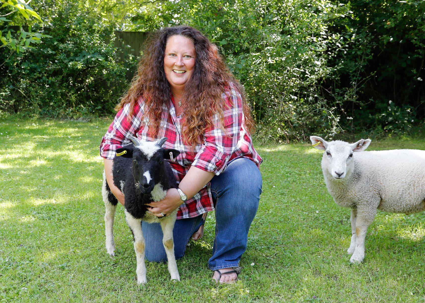 Beth crouching down on grass with her arms around one sheep and another sheep on her right hand side