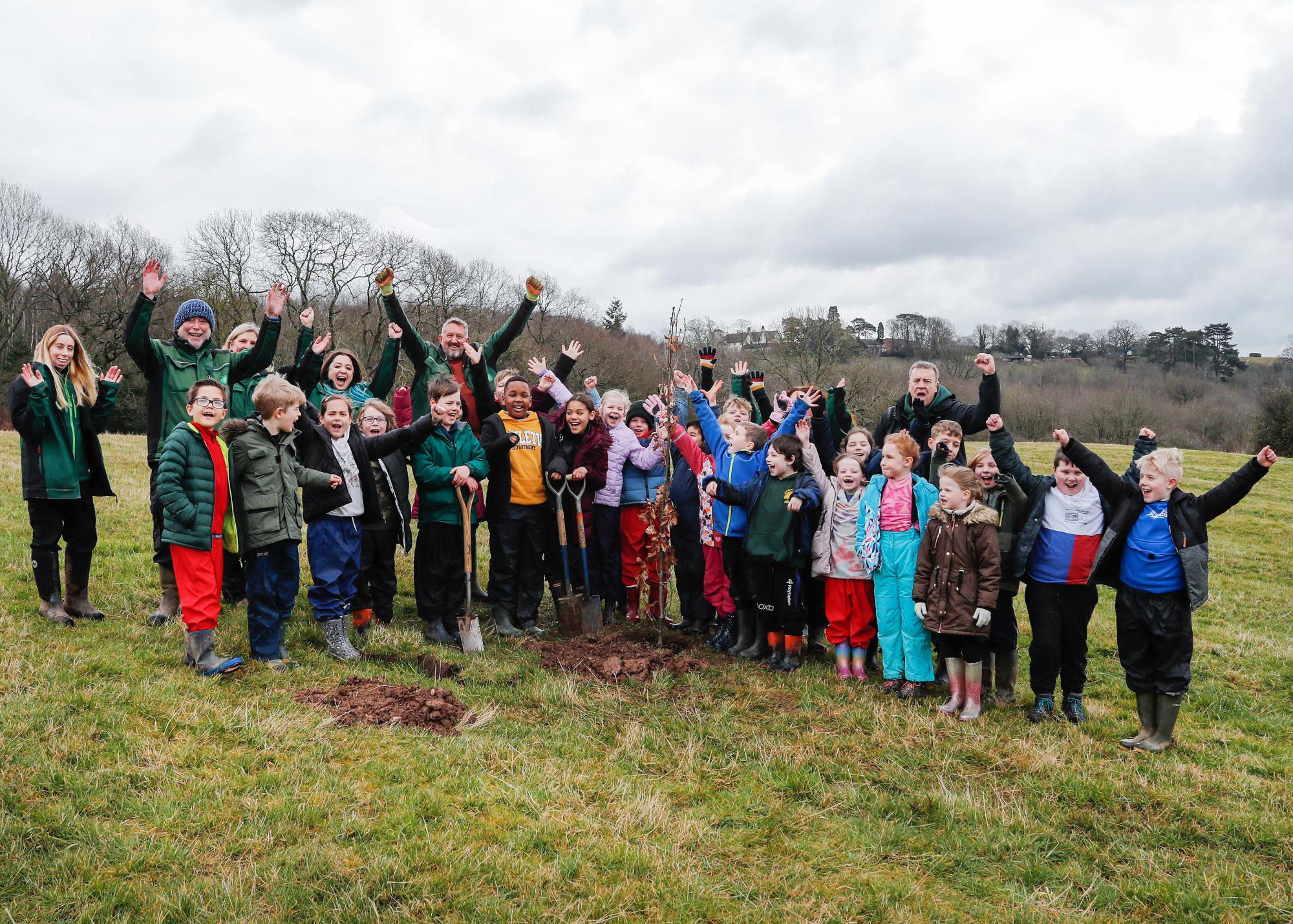 Large group of school pupils with school staff and members of the Forest team cheering with their hands in the air, standing behind the newly planted 2 millionth tree