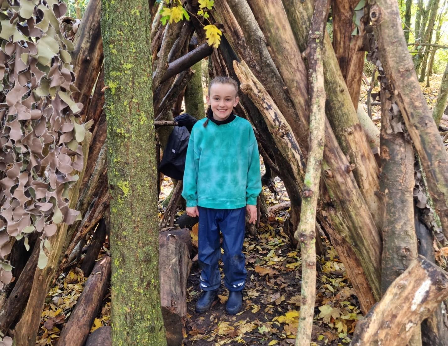 A young girl standing in a den made of tree branches in a woodland clearing smiling at the camera