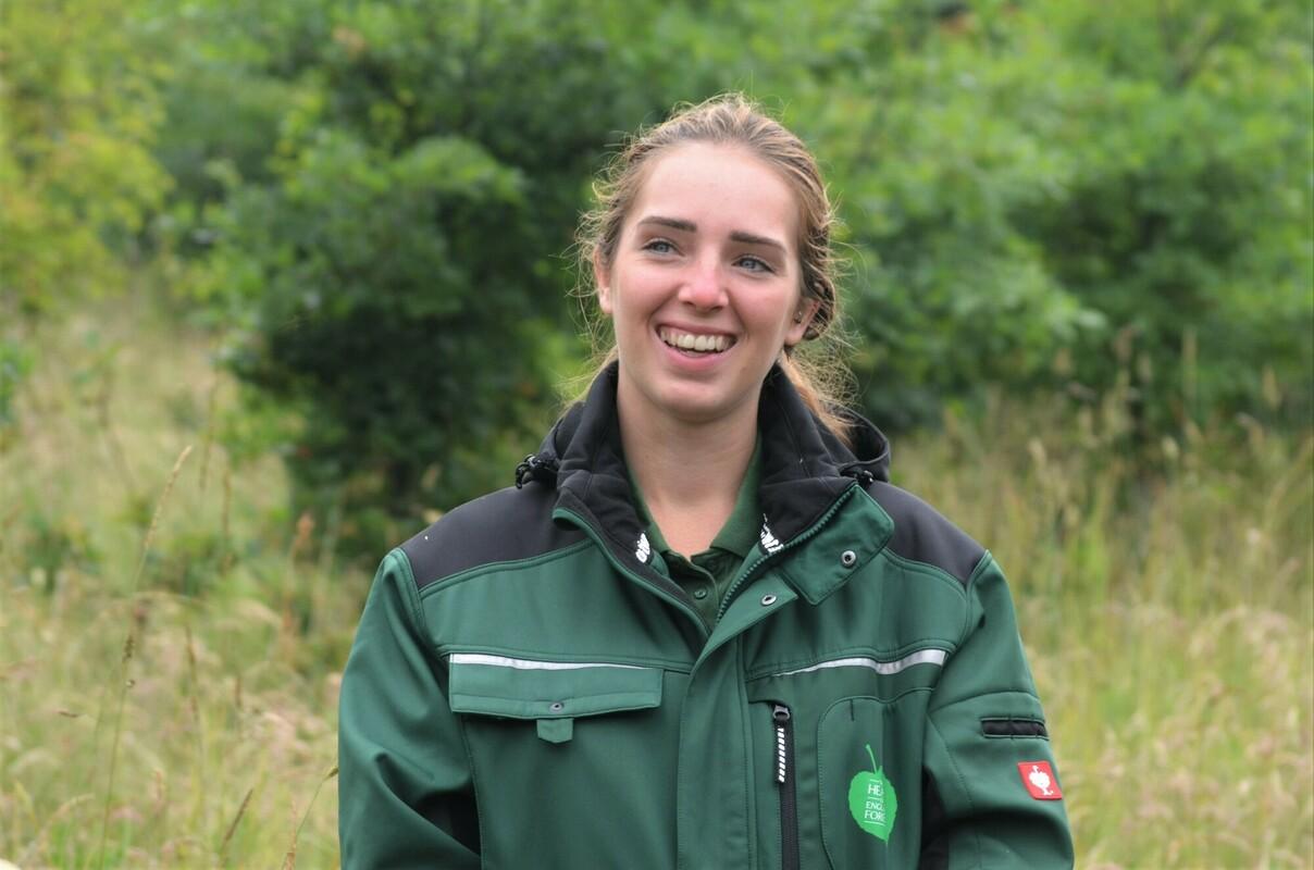 Emma, our Assistant Biodiversity Officer (Grasslands) standing in the Forest wearing a charity branded coat. She is smiling at the camera