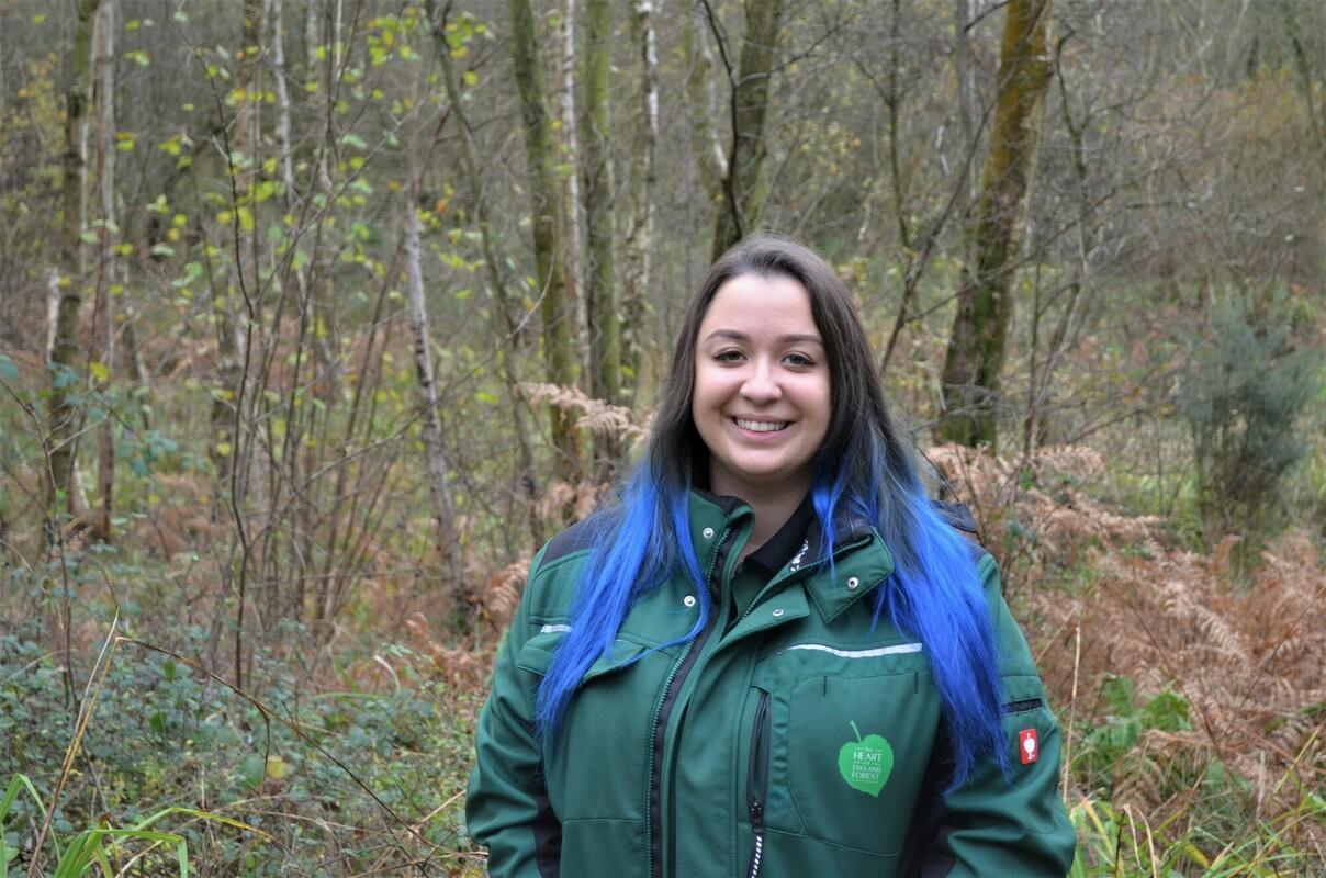 Gorcott Biodiversity Officer, Tasha, looking at the camera smiling with woodland behind her