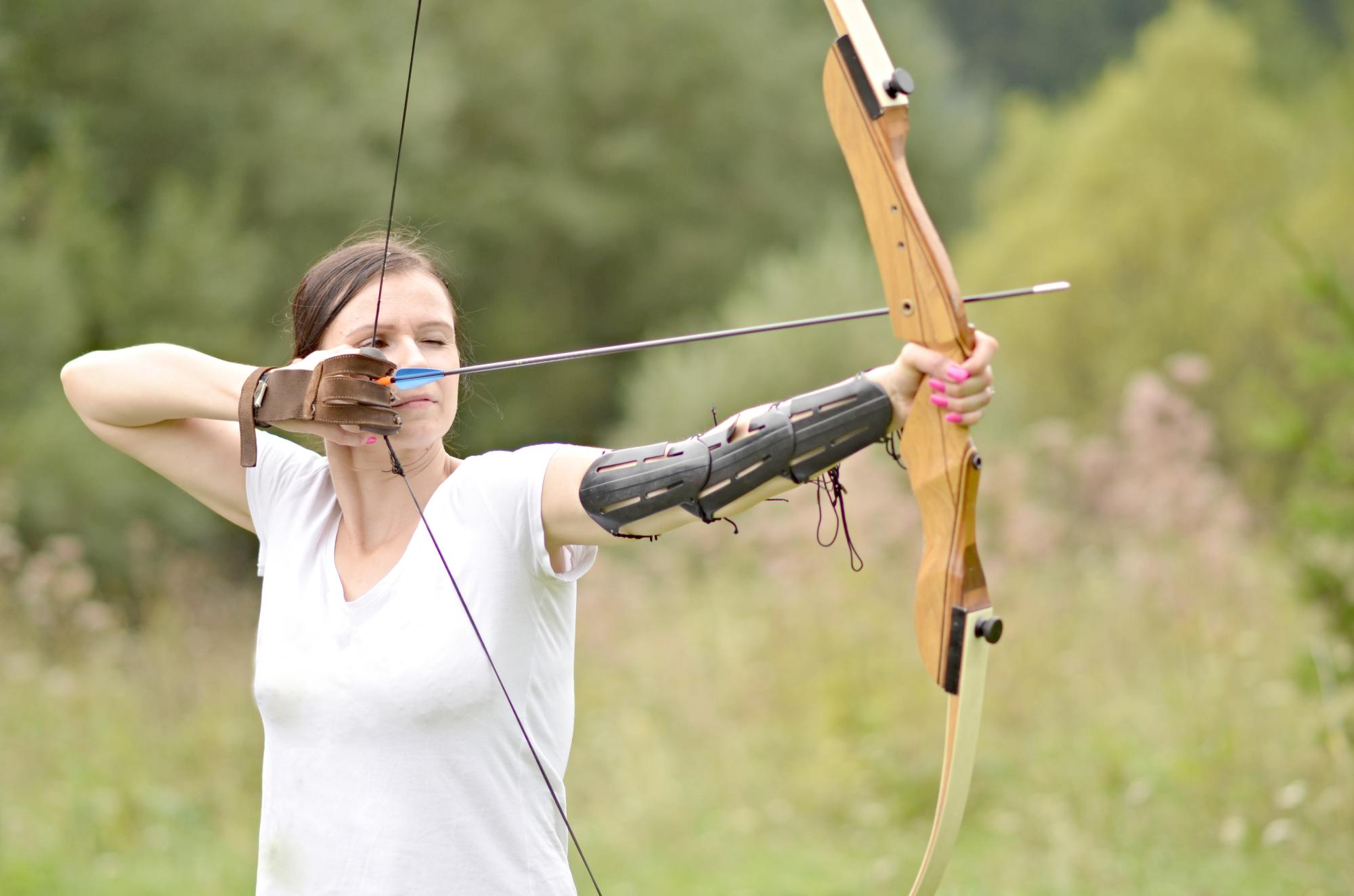 A women holding a bow, pulling back the string about to release and arrow. She is wearing the appropriate safety gear. Shutterstock.