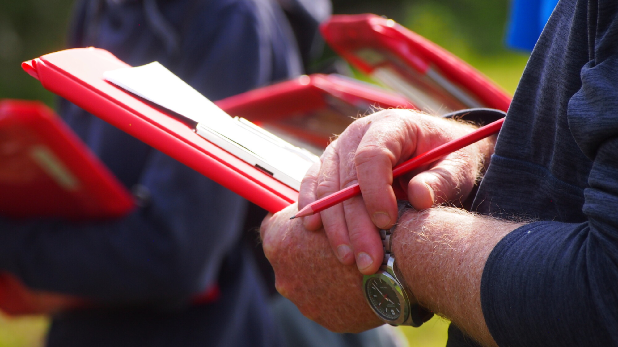 A close up shot of a hand holding a red clipboard with a pencil in the other hand