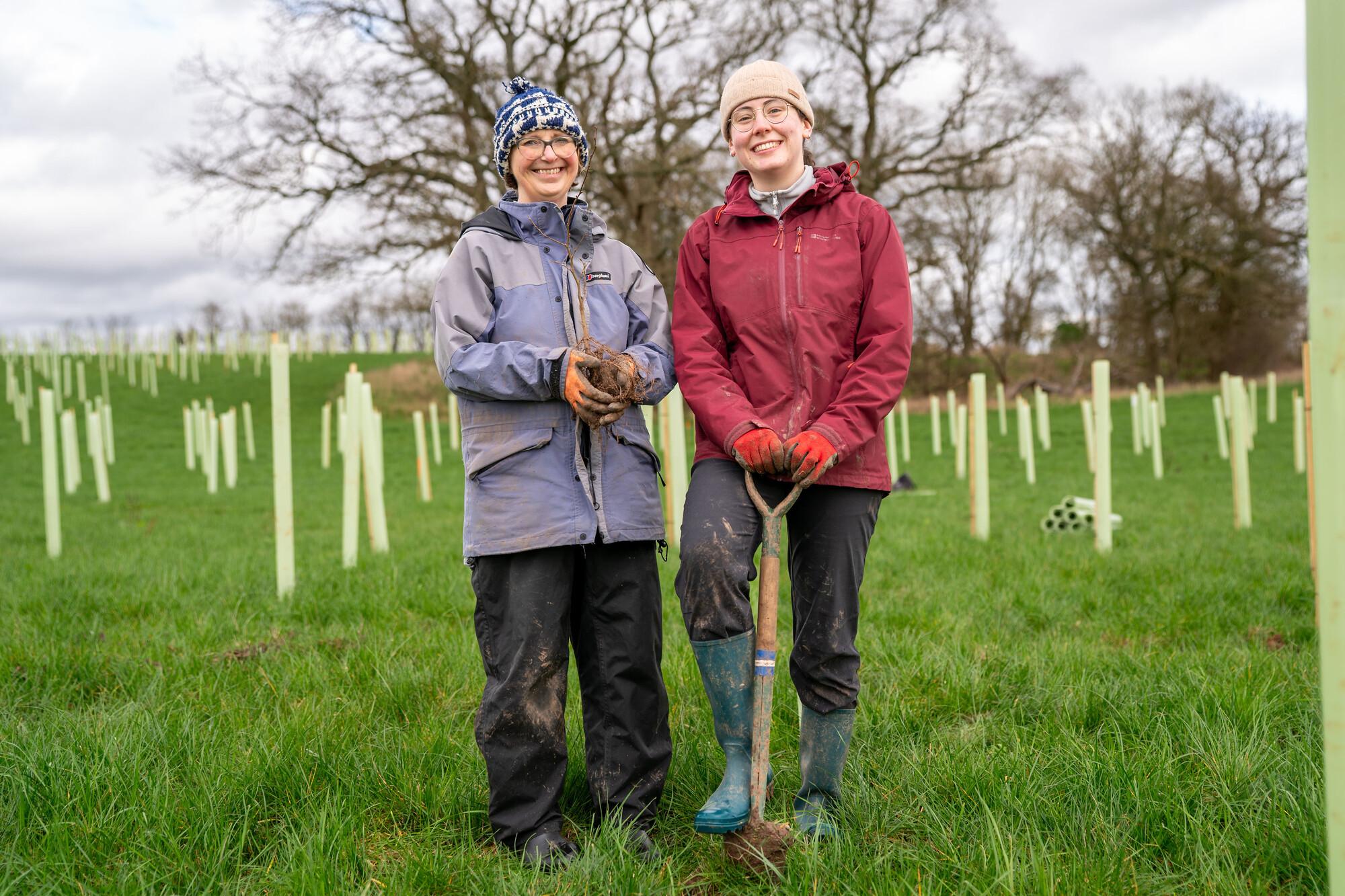 Two female volunteers - one holding a tree sapling and one resting her hands on a spade, during a tree planting session at Eastern Hill