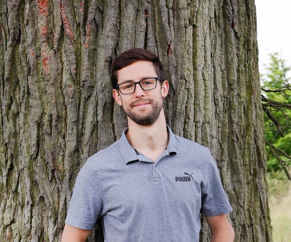 Alexander leaning on a tree trunk, smiling at the camera.