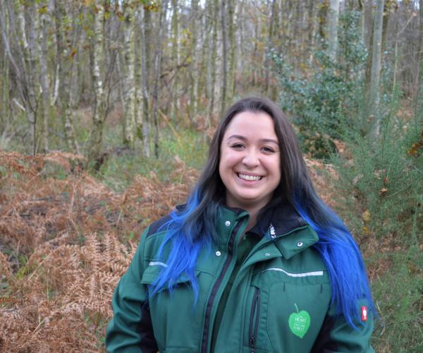 Head and shoulders shot of Tasha smiling at the camera wearing a branded jacket and standing in front of some autumnal coloured scrub