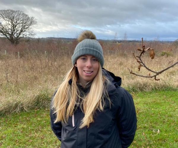 Close up of Ellie smiling in a fluffy grey beanie hat stood on some grass in the Forest.